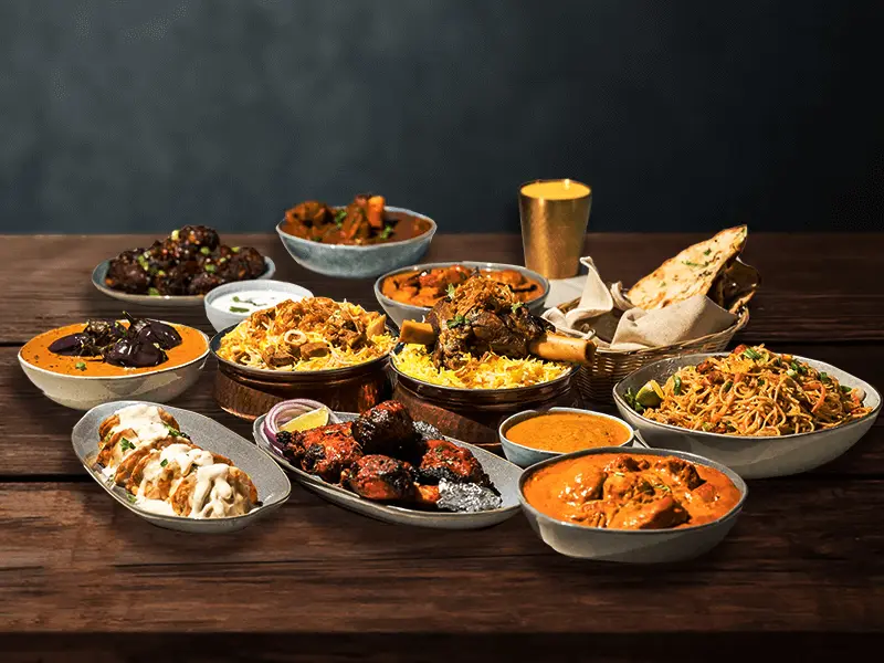 A full table of Kachiguda Junction dishes — mutton shank biryani, butter chicken, baingan curry, tandoori chicken, naan and noodles on dark wood